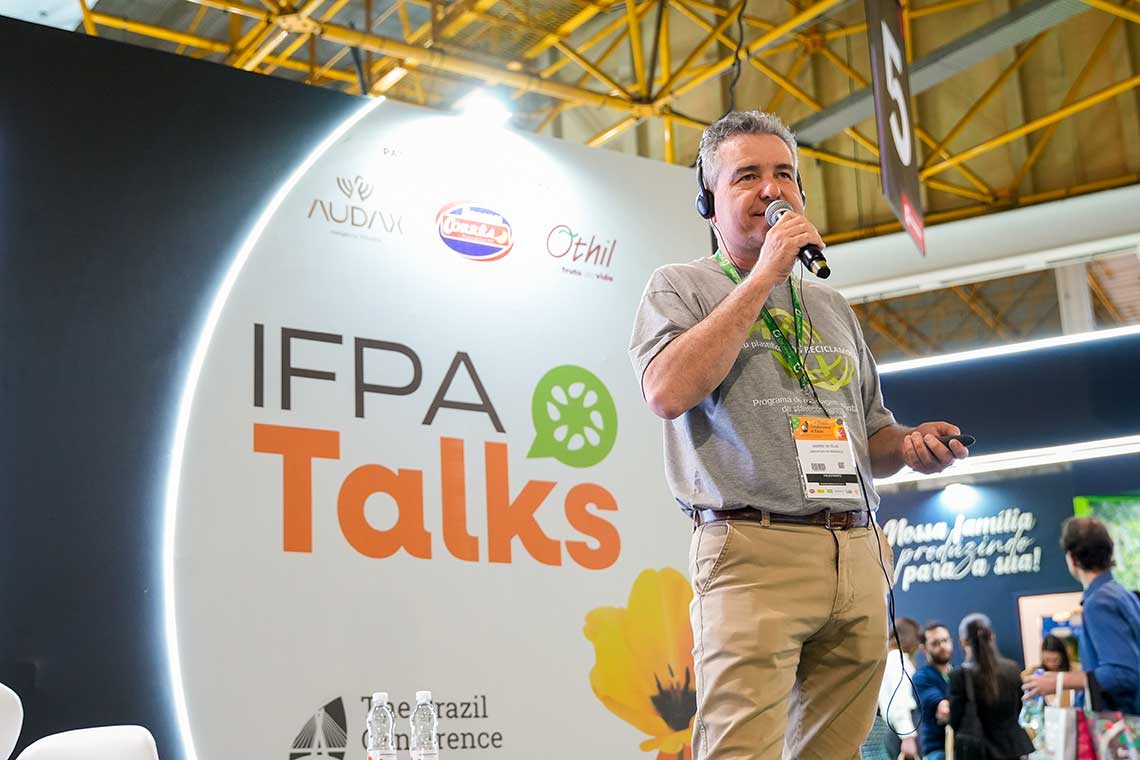 A man holding a microphone is speaking on a stage at the IFPA Talks event. He is wearing a gray t-shirt, beige pants, and a lanyard with a badge. Behind him is a large IFPA Talks sign along with logos and some attendees are visible in the background.