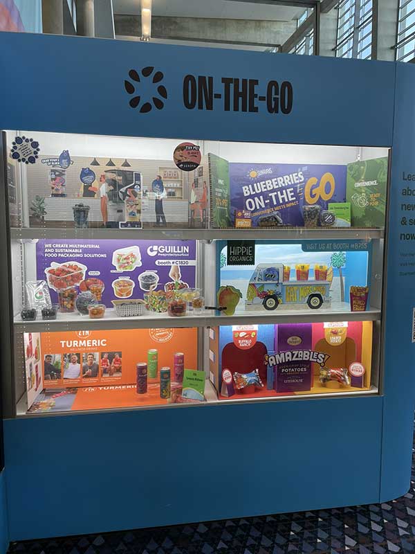 An exhibition display case titled "ON-THE-GO," showcasing blueberries, sustainable packaging, and turmeric and potato products.