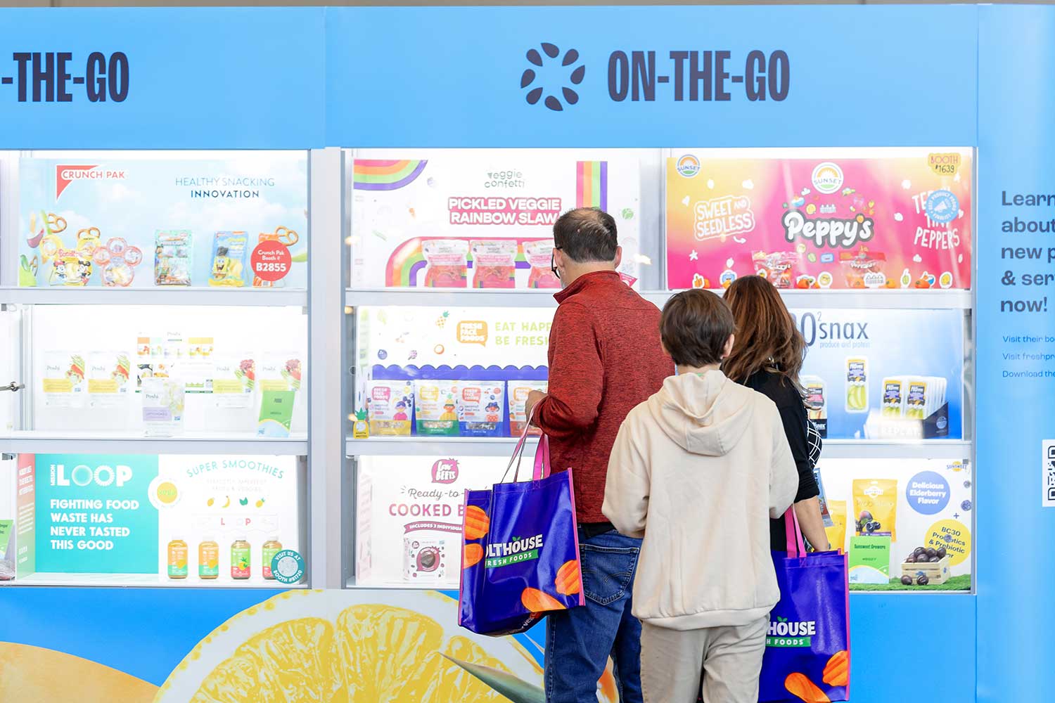 People viewing a snack display labeled "ON-THE-GO" with various colorful packaged snacks.