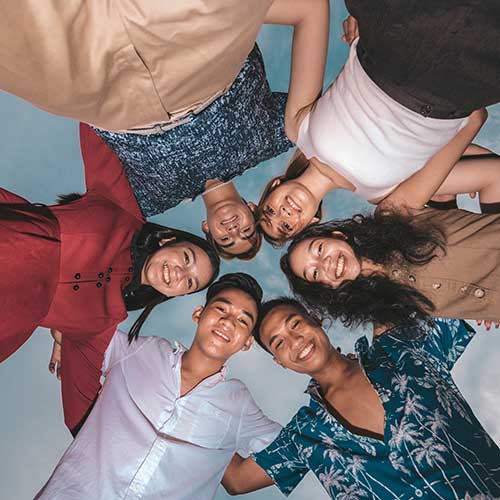 A circle of six young friends posing happily for the camera. Shot from the ground looking up. 