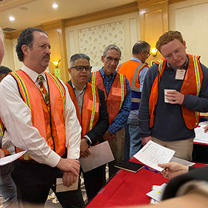 A group of people wearing orange safety vests in a meeting, holding papers in a brightly lit room.